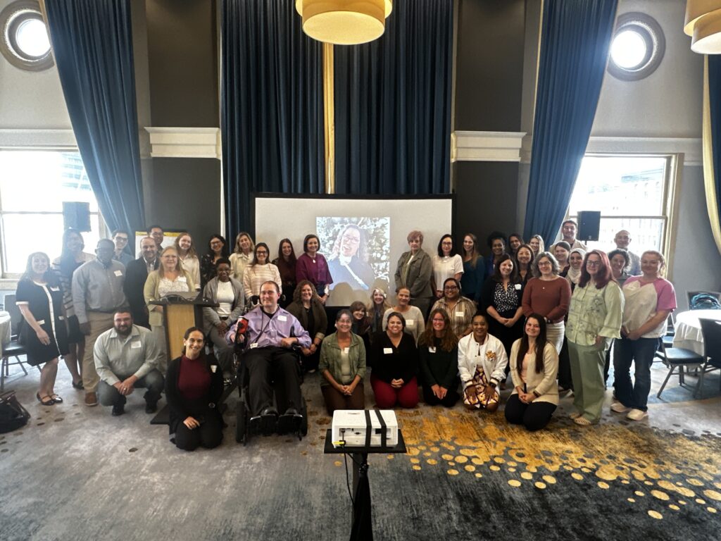 NCI-IDD 2026 Annual Meeting attendees gathered around a photo of Caitlin Bartley